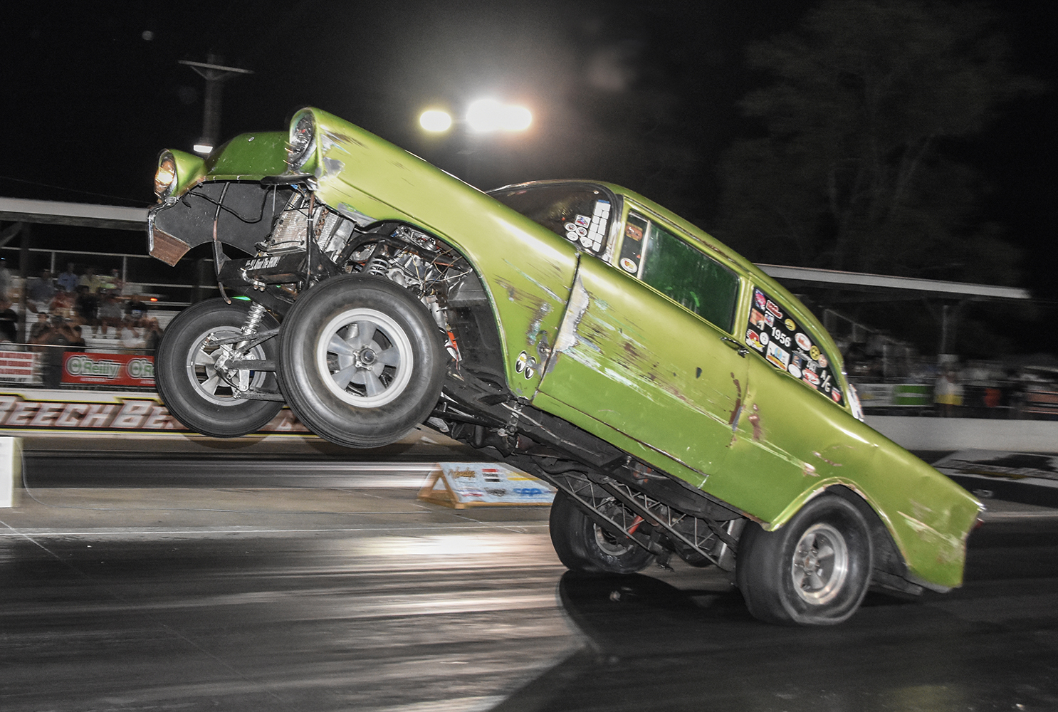 03 1956 Chevrolet drag car launching on the strip with a distressed green body and high wheelie stance under night lights
