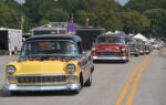 12 1956 Chevrolet 210 in black and yellow with flame graphics leading a line of Tri Five Chevys in parade