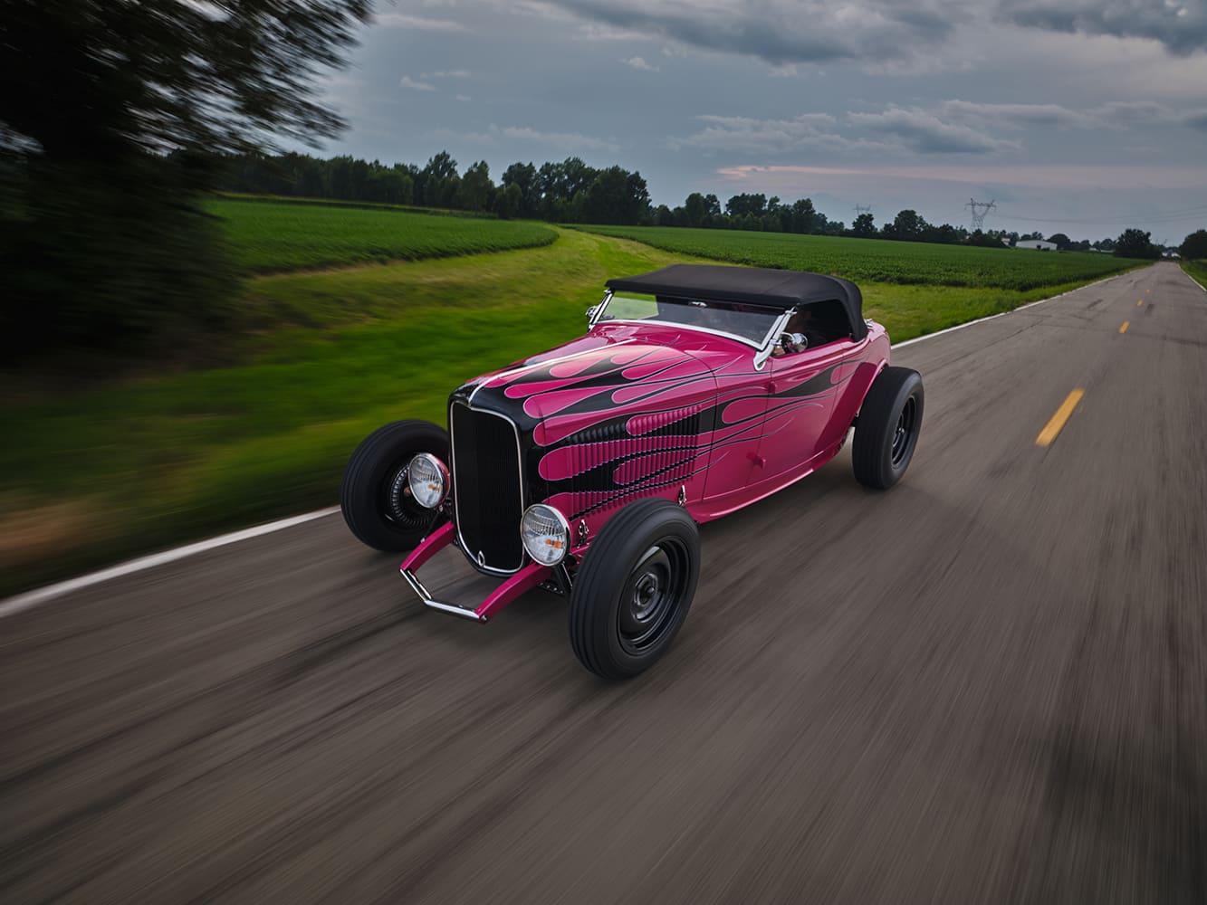 12 1932 Ford Highboy Roadster with black soft top and magenta flame job cruising a scenic rural highway