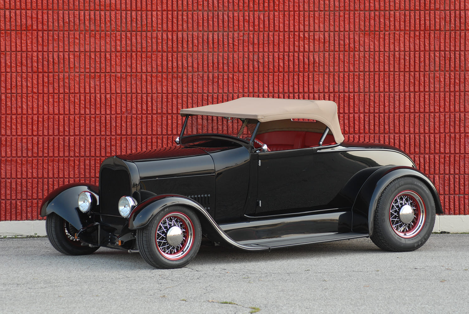 30 1930 Ford Roadster with tan top red accents and wire wheels against a textured red brick backdrop