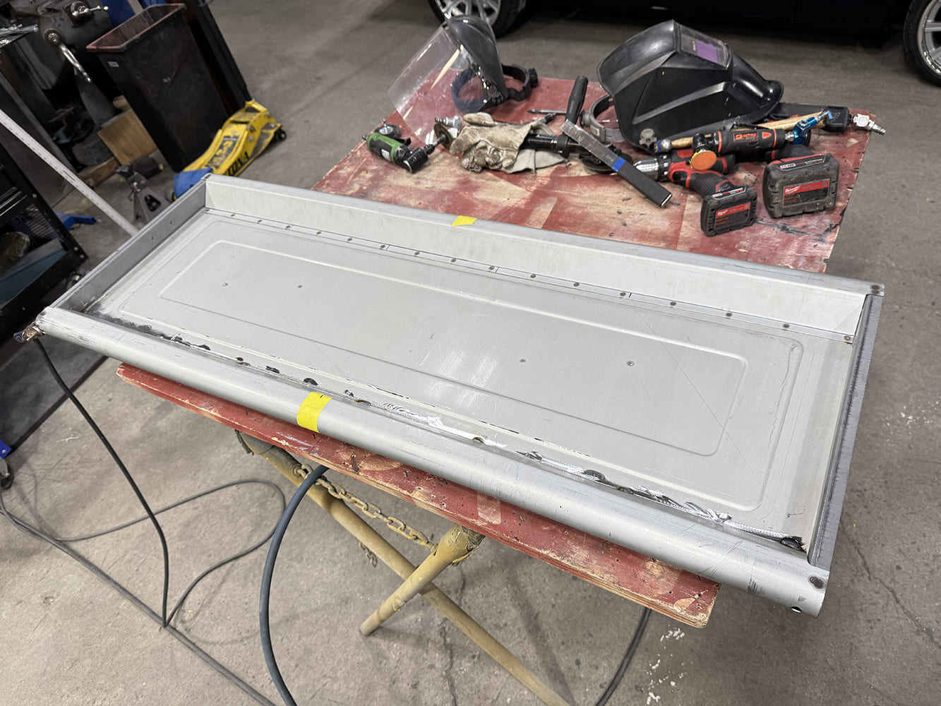 02 Tailgate on reddish worktable perimeter weld seams visible tools scattered concrete floor vehicle tires behind