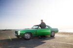 015 1969 Chevrolet Camaro SS green coupe beachside photo with owner standing by driver door in sunglasses and cap