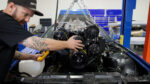 Mechanic guiding a high-performance V8 engine with black serpentine pulley system into a blue third-generation Chevy Camaro engine bay using an engine hoist.