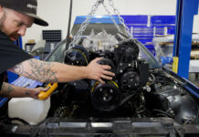 Mechanic guiding a high-performance V8 engine with black serpentine pulley system into a blue third-generation Chevy Camaro engine bay using an engine hoist.