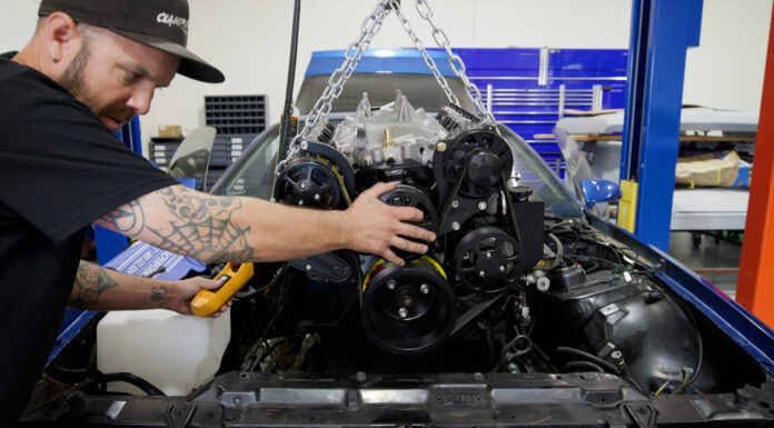Mechanic guiding a high-performance V8 engine with black serpentine pulley system into a blue third-generation Chevy Camaro engine bay using an engine hoist.