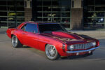 Front three-quarter view of a custom candy red metallic 1969 Chevy Camaro coupe featuring a high-rise cowl induction hood, polished multi-spoke wheels, and modern pro-touring stance parked outdoors.