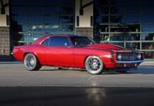 Low-angle side profile view of a candy apple red metallic 1969 Chevy Camaro restomod parked on concrete, featuring a cowl induction hood, polished deep-dish wheels, and tinted windows.
