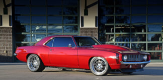 Low-angle side profile view of a candy apple red metallic 1969 Chevy Camaro restomod parked on concrete, featuring a cowl induction hood, polished deep-dish wheels, and tinted windows.