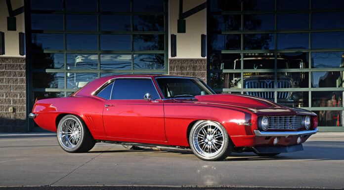 Low-angle side profile view of a candy apple red metallic 1969 Chevy Camaro restomod parked on concrete, featuring a cowl induction hood, polished deep-dish wheels, and tinted windows.