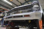 Low-angle front view of a second-generation Chevy Nova featuring a custom-fabricated, brushed steel reshaped front bumper and lower valance with integrated turn signals in a shop.