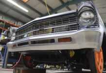 Low-angle front view of a second-generation Chevy Nova featuring a custom-fabricated, brushed steel reshaped front bumper and lower valance with integrated turn signals in a shop.