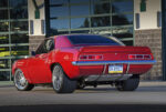 Rear three-quarter view of a custom metallic red 1969 Chevy Camaro restomod showing wide rear performance tires, rear spoiler, chrome bumper, and custom taillight panel.