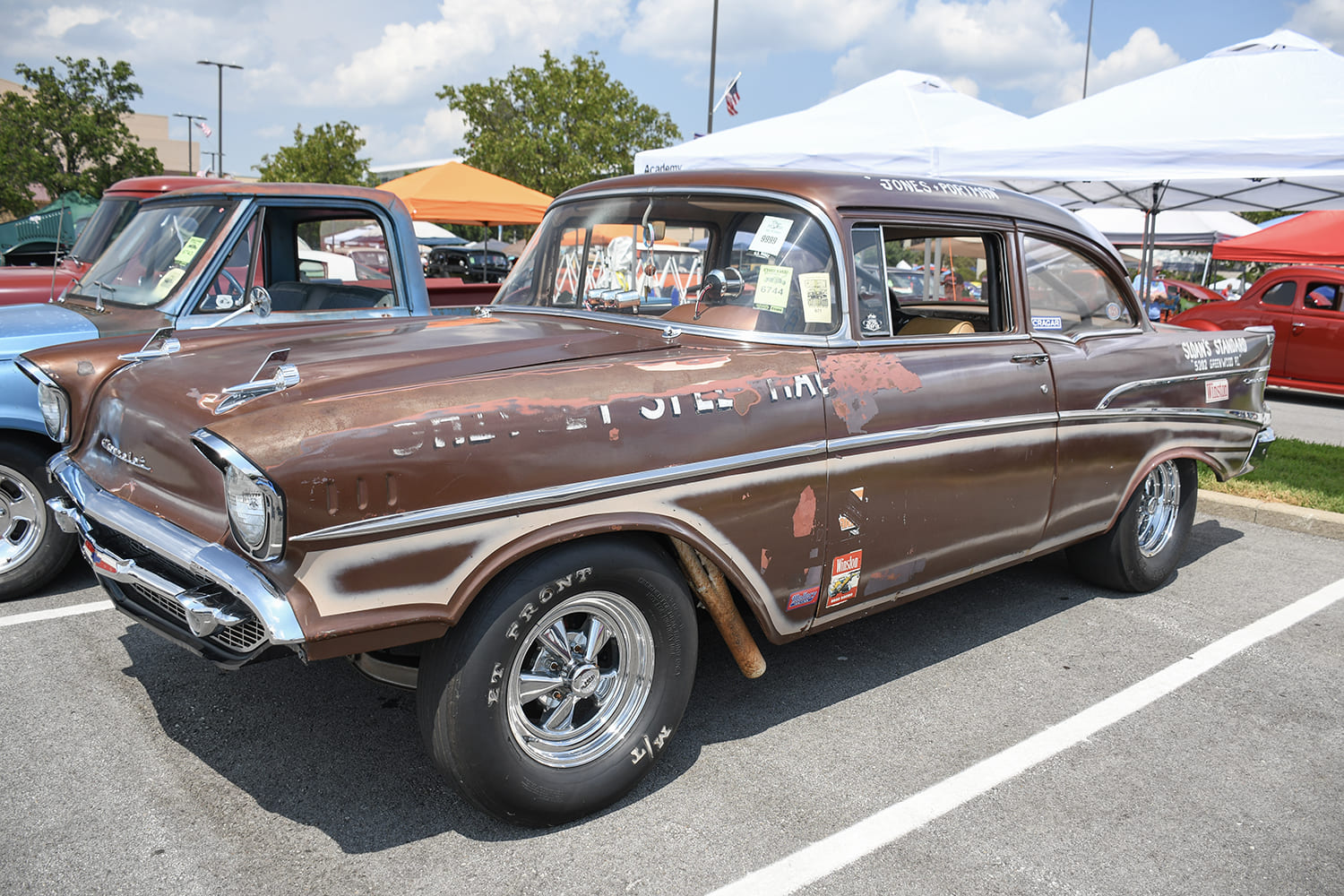Front quarter shot of a vintage brown 1957 Chevy two-door post gasser drag car with weathered patina and open fender-well headers.