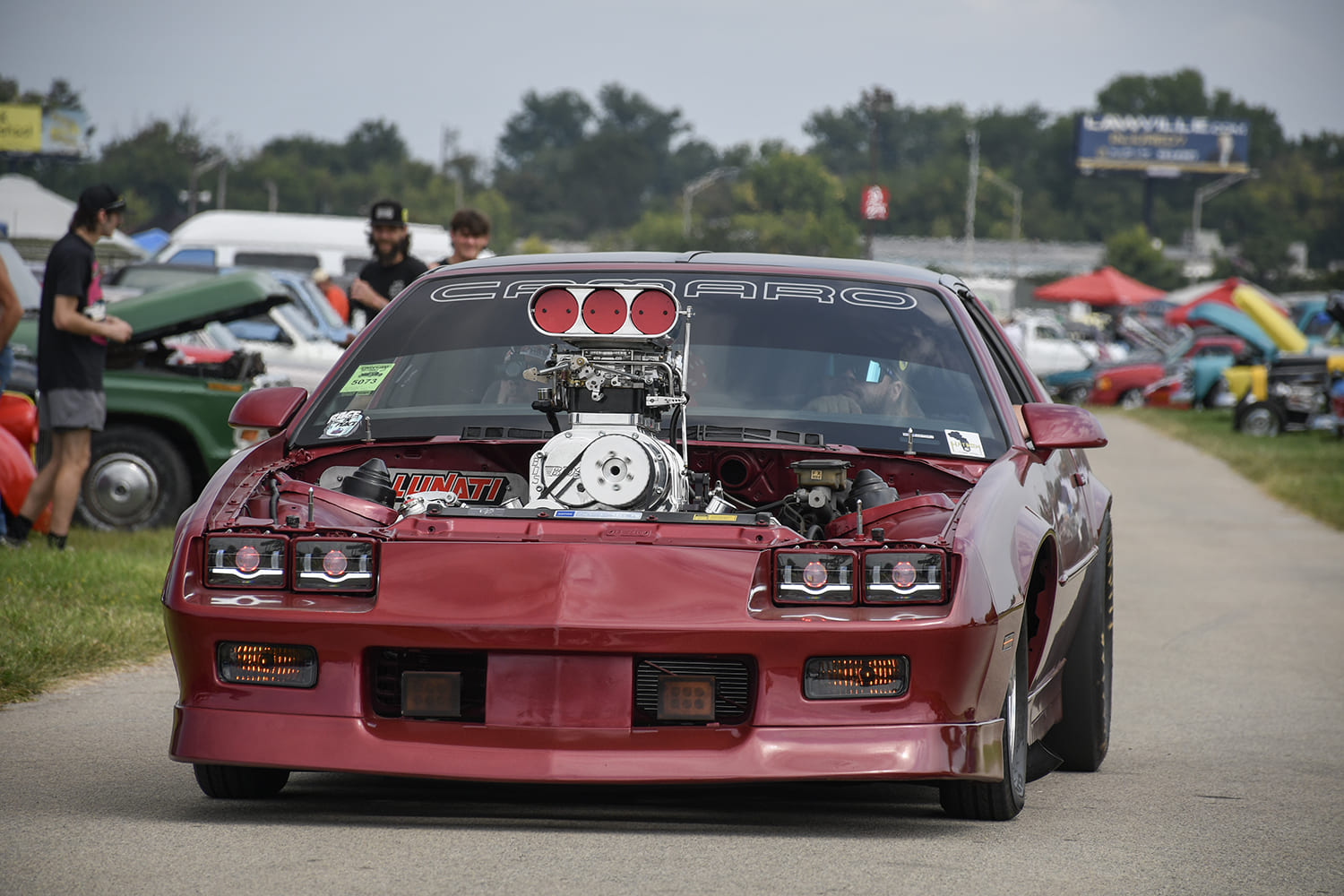 Front view of a maroon third-generation Chevrolet Camaro driving with an exposed supercharged engine and hood removed at the NSRA Street Rod Nationals.