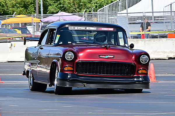 Front three-quarter view of a dark red and black 1955 Chevrolet Tri-Five autocross race car driving on the track with an aero rear wing at the Goodguys Pleasanton West Coast Nationals.