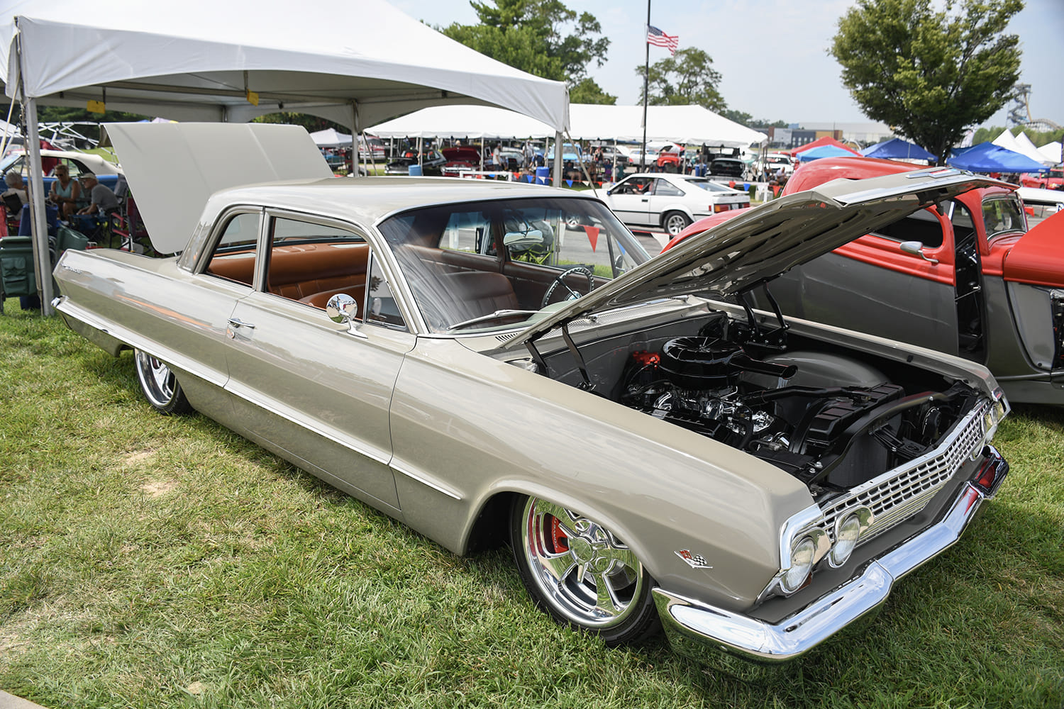 Front three-quarter view of a lowered champagne-silver 1963 Chevy Impala muscle car with an open hood, custom chrome wheels, and tan interior on a show lawn.