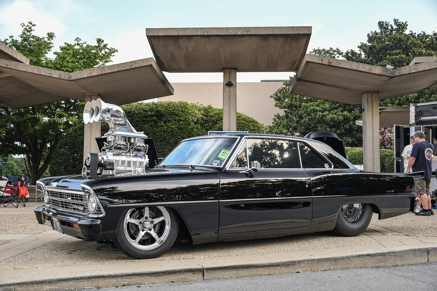 Side profile of a gloss black second-generation Chevy II Nova street machine featuring a huge polished supercharger scoop towering over the hood and wide rear slicks.