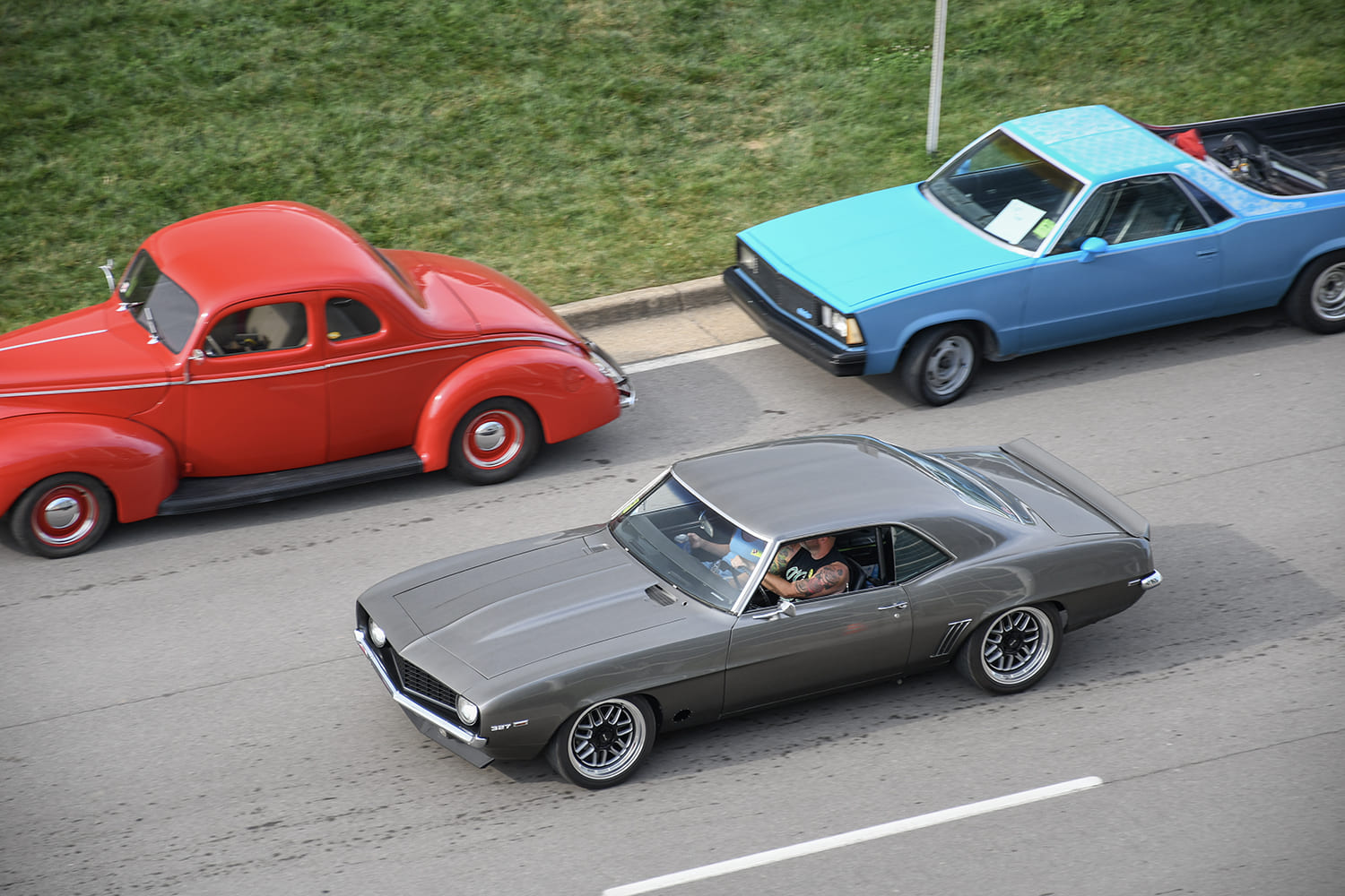 High-angle overhead shot of a custom gunmetal grey 1969 Chevrolet Camaro driving on the road flanked by a red vintage coupe and a blue pickup.