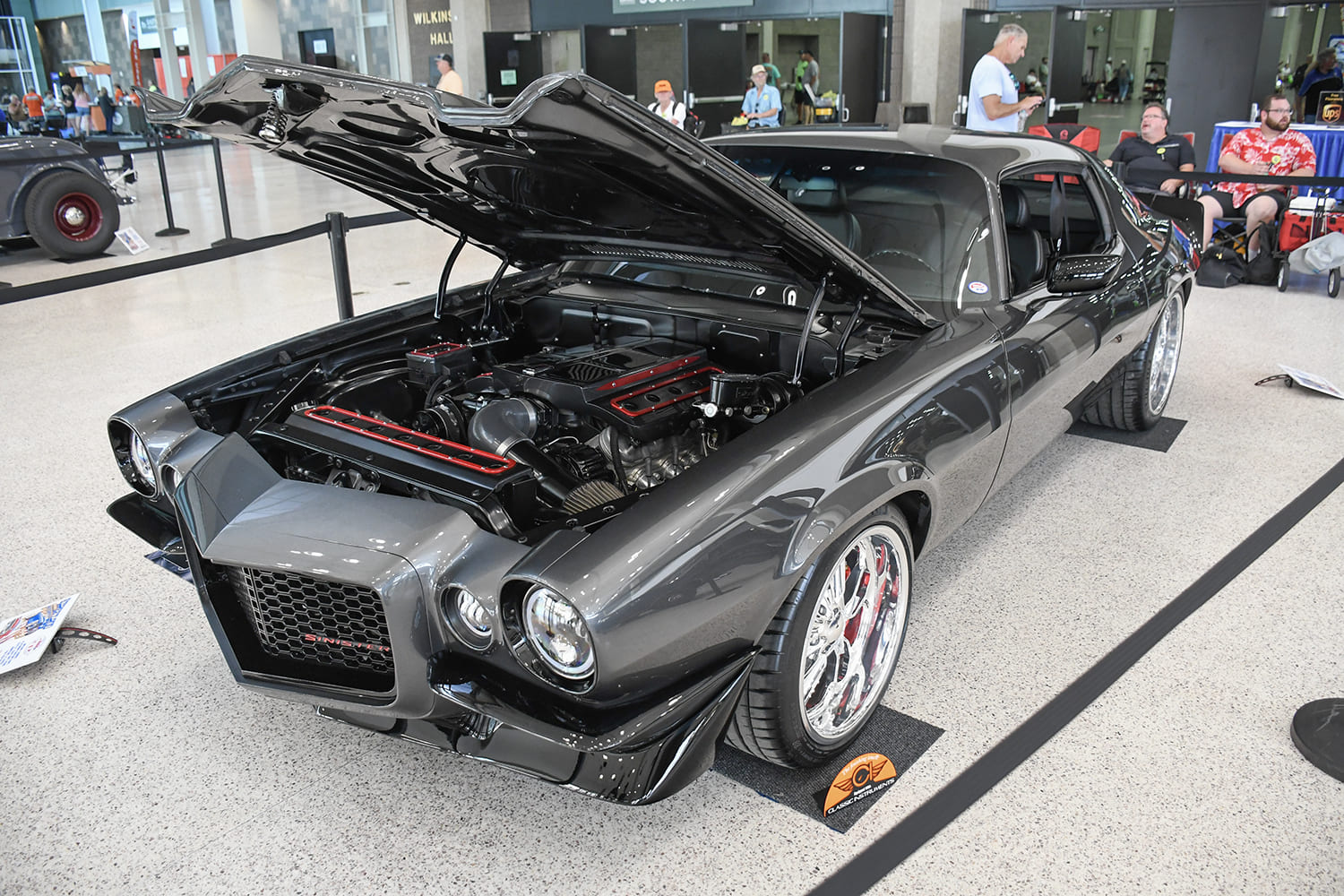 High-angle indoor shot of a metallic charcoal split-bumper second-generation Chevy Camaro with an open hood revealing a modern LS engine with red accents.