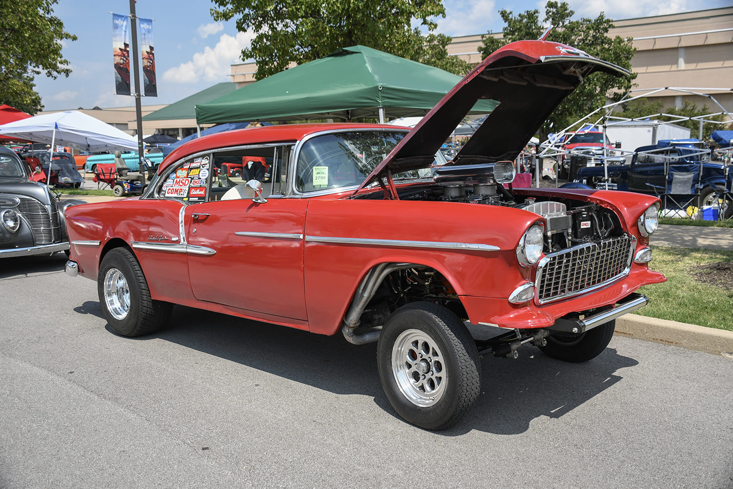High-riding red 1955 Chevrolet Bel Air gasser with straight front axle, raised tilt hood, chrome tunnel ram scoop, and vintage race decals.