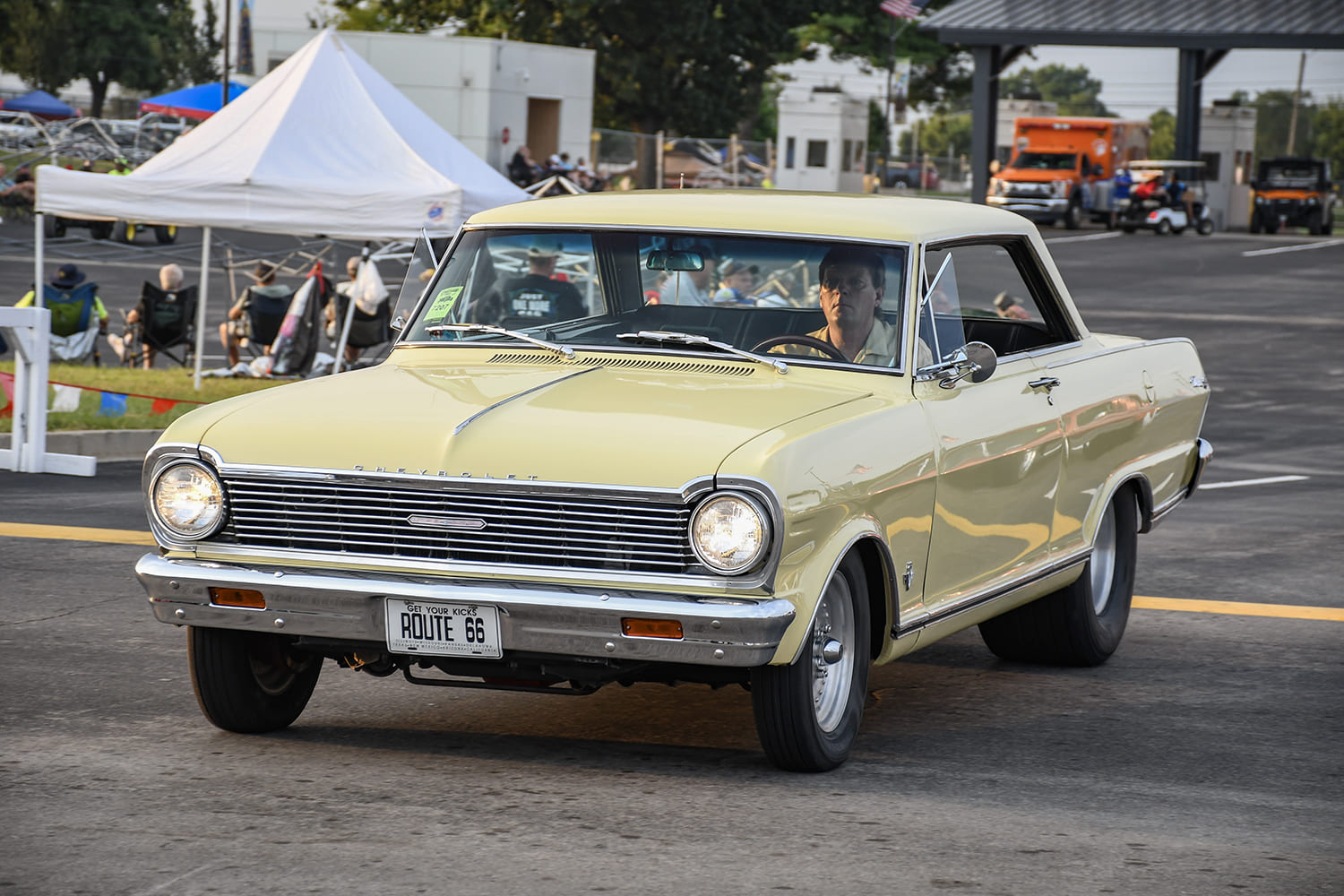 Front three-quarter view of a pale yellow first-generation Chevrolet Nova hardtop cruising on the pavement with classic dish wheels.