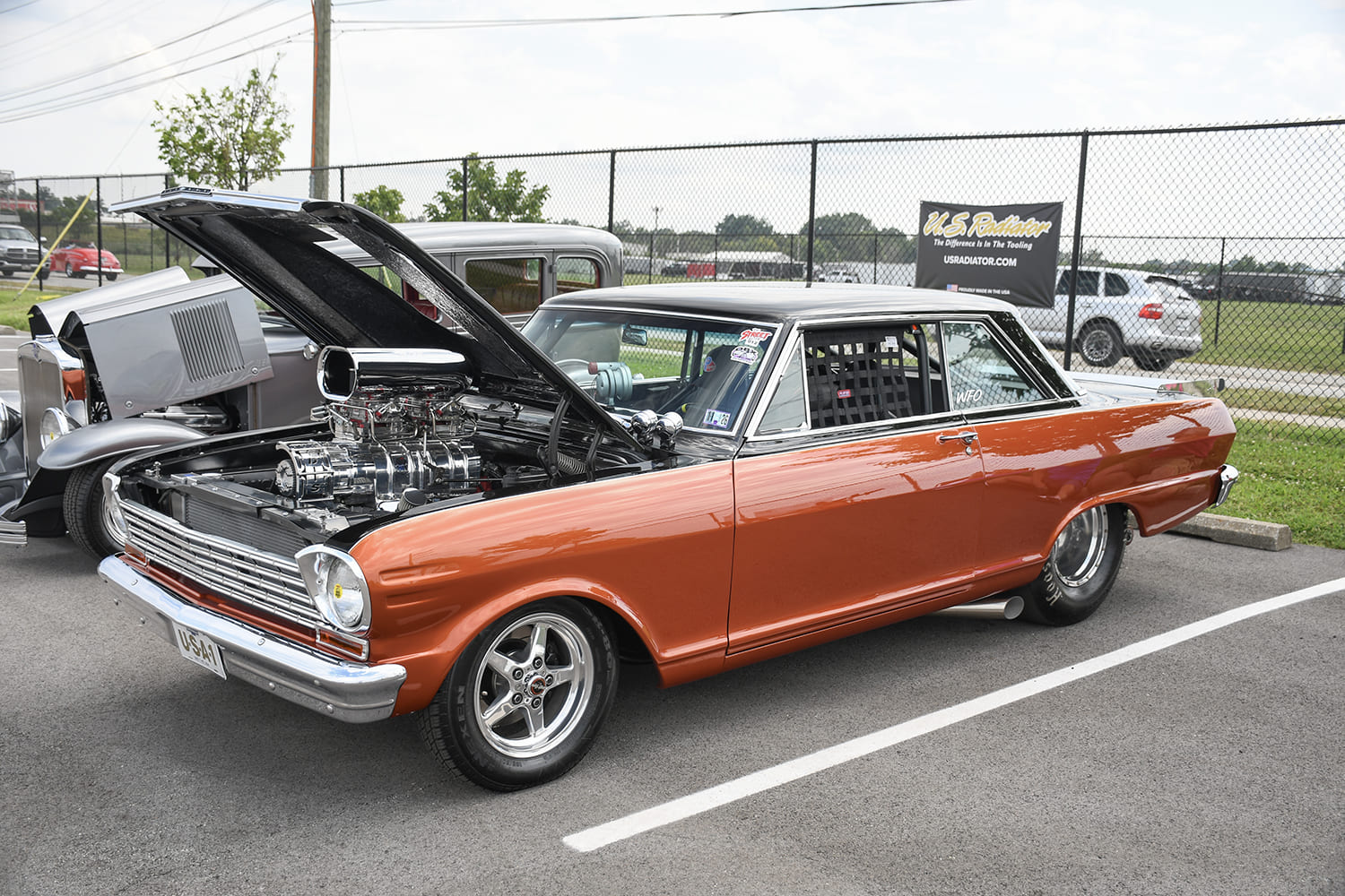 Copper-orange and black two-tone classic Chevy II Nova drag car with an open hood showcasing a polished supercharger and chrome intake scoop.