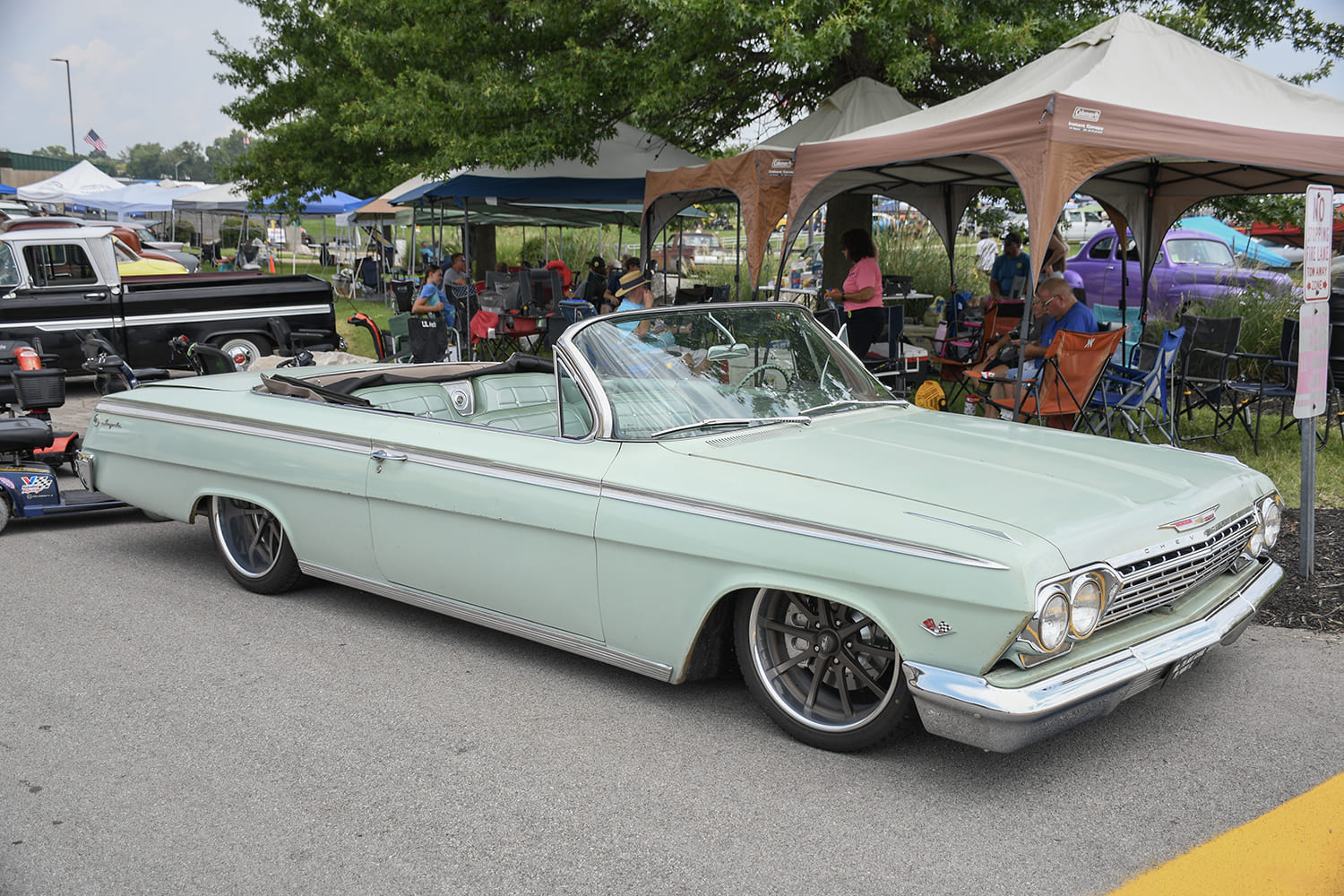 Front three-quarter profile of a slammed seafoam green 1962 Chevrolet Muscle Car convertible with large modern wheels parked under canopies.