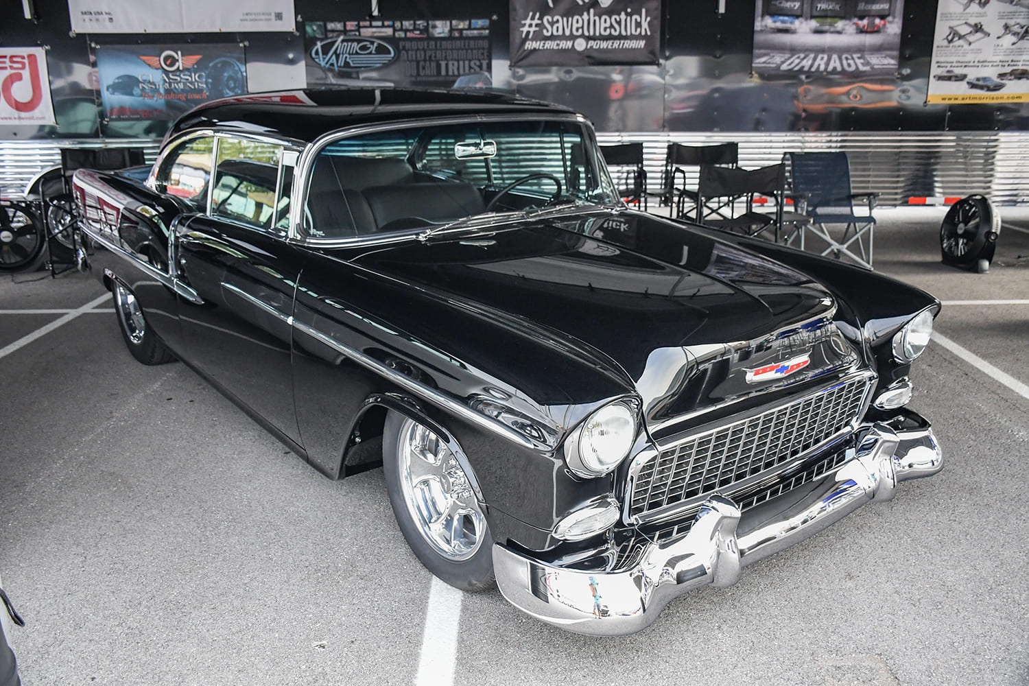 Front quarter view of a slammed gloss black 1955 Chevrolet Bel Air hardtop with chrome multi-spoke wheels parked at a vendor display.