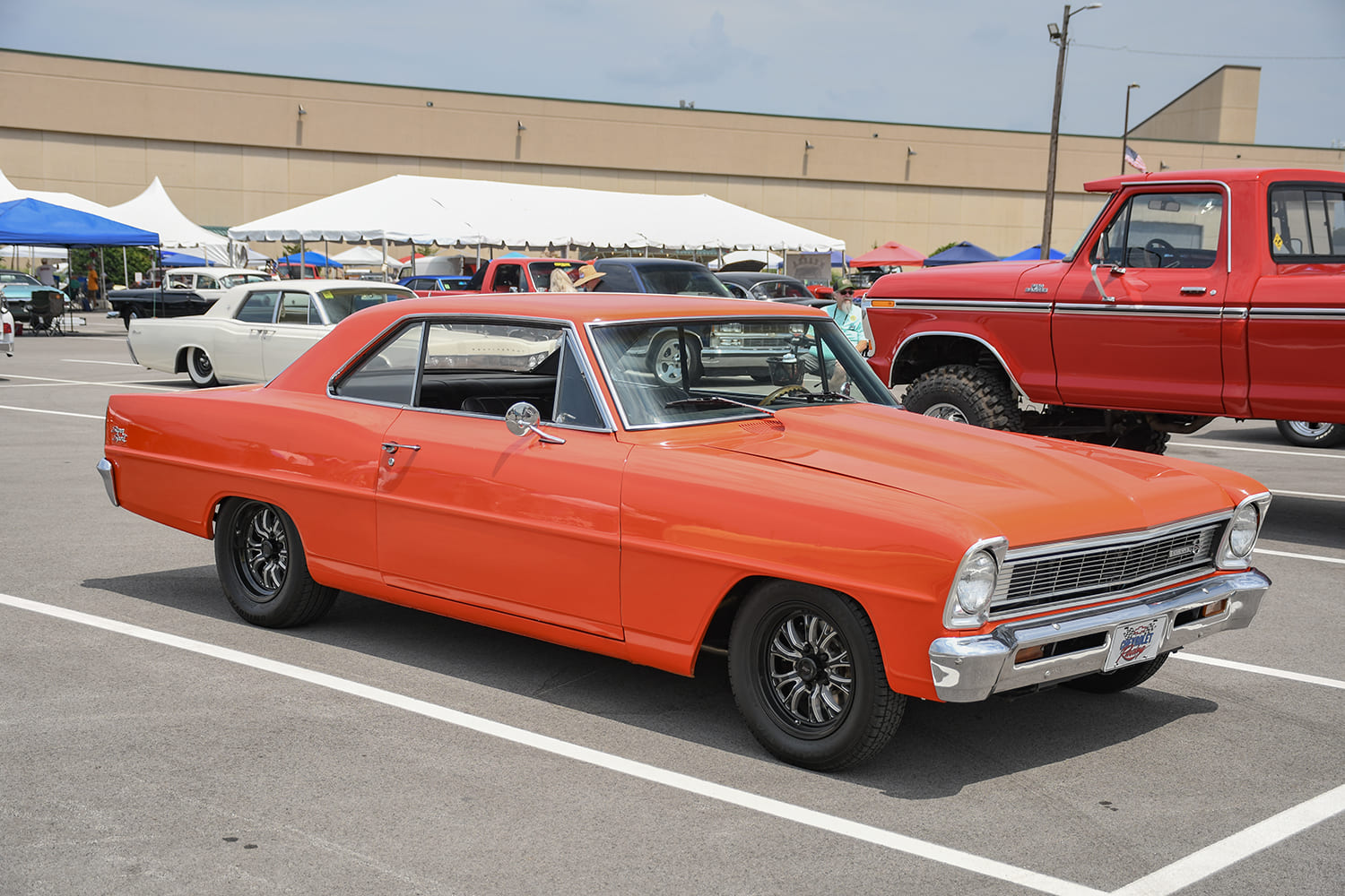 Front quarter profile of a bright orange 1966–1967 Chevrolet Chevy II Nova coupe with black multi-spoke wheels parked in an outdoor lot.