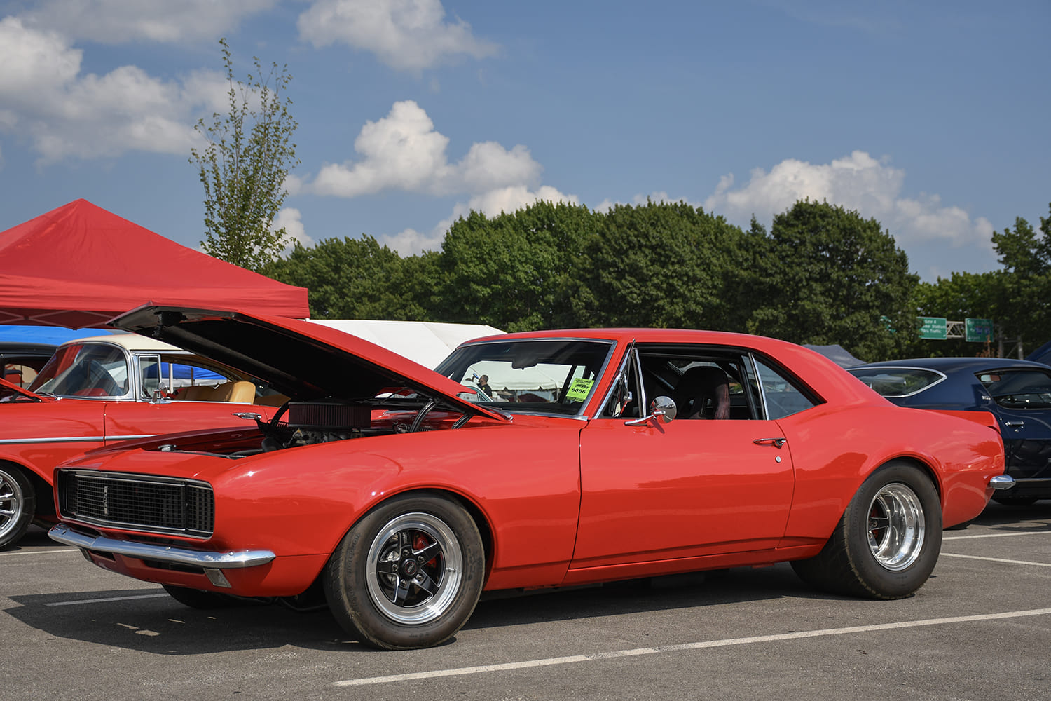 Front three-quarter view of a vibrant red first-generation Chevy Camaro RS with an open hood, black-center wheels, and massive rear drag slicks.