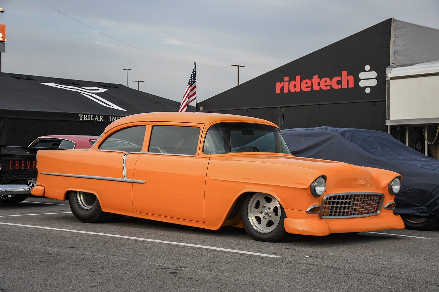Side profile of a low-riding pastel orange 1955 Chevy two-door post sedan with billet wheels and wide rear slicks parked at an outdoor event.