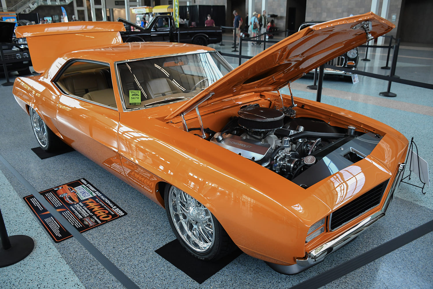 Front three-quarter view of a custom candy orange 1969 Chevrolet Camaro restomod with an open hood displaying a detailed V8 engine and polished wheels indoors.