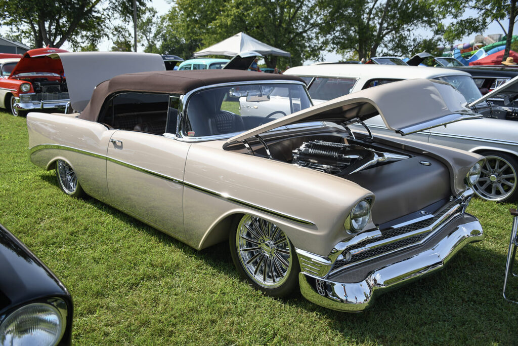 Front quarter shot of a champagne metallic 1956 Chevrolet Bel Air convertible with a brown top, custom wire-style chrome wheels, and an open hood revealing a modern supercharger.
