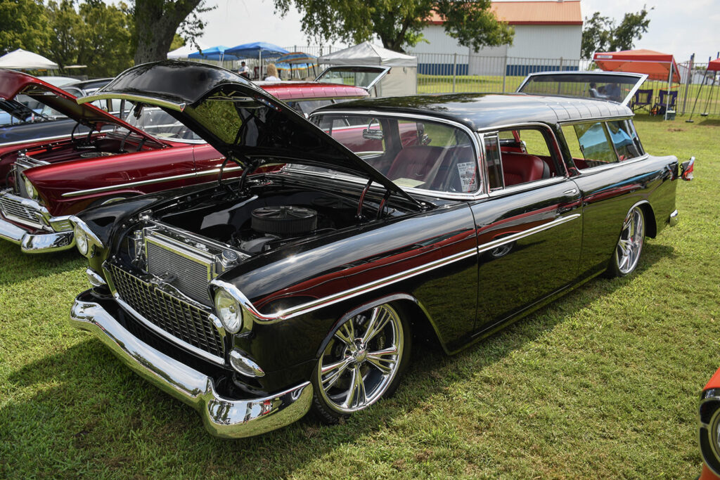 High-angle front three-quarter view of a custom gloss black 1955 Chevrolet Nomad wagon with polished multi-spoke wheels and an open engine bay on the grass.