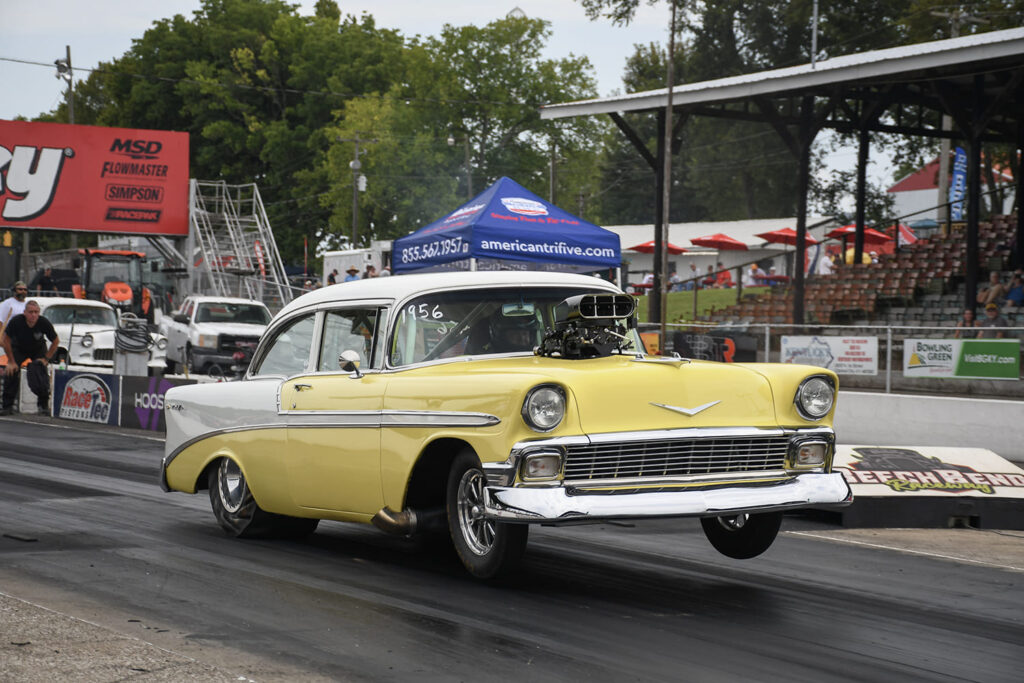 Yellow and white two-tone 1956 Chevrolet Bel Air drag car with an exposed supercharger scoop lifting its front wheels on the drag strip at the Tri-Five Nationals.