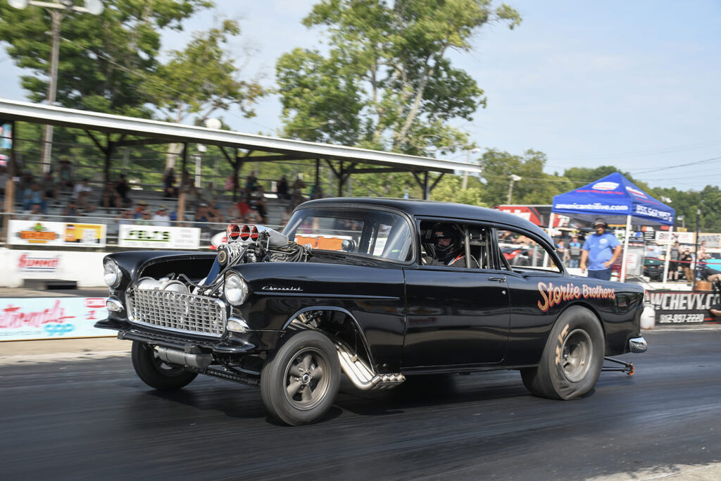 Side profile action shot of a black 1955 Chevrolet gasser with a tall supercharger scoop and fender-well headers speeding down the Beech Bend drag strip.