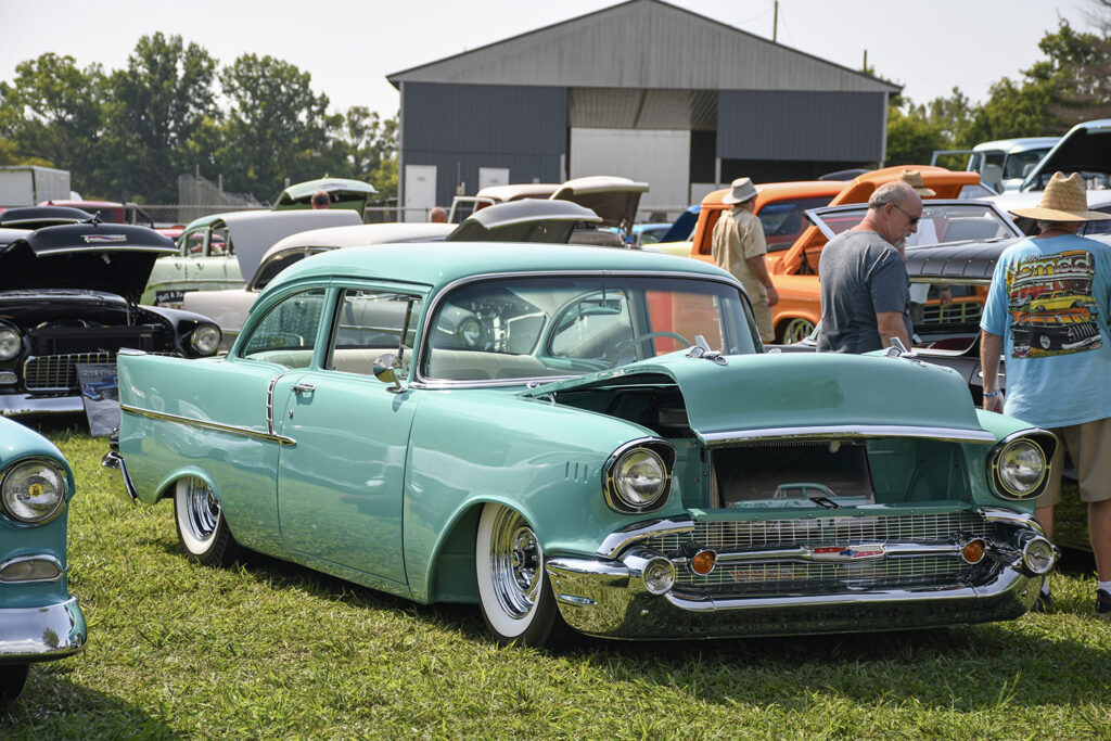 Front three-quarter view of a lowered green 1957 Chevrolet Bel Air two-door post with chrome smoothie wheels and whitewall tires parked on the lawn.