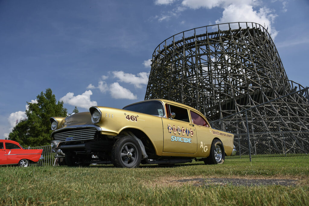 Low-angle front quarter view of a gold 1957 Chevrolet gasser race car parked on the grass in front of the Beech Bend wooden roller coaster.