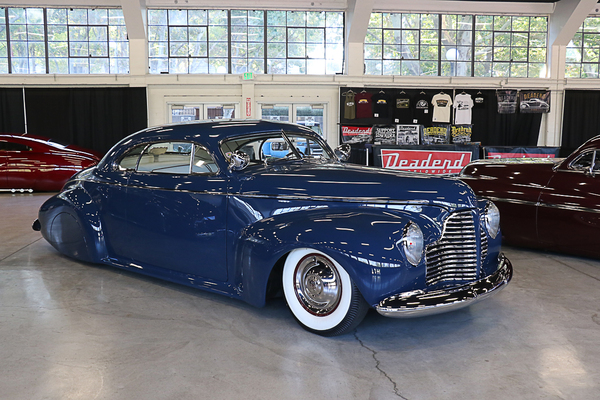 Front three-quarter view of a dark blue chopped 1940 Buick custom coupe with wide whitewall tires on display indoors at the Goodguys Pleasanton West Coast Nationals.