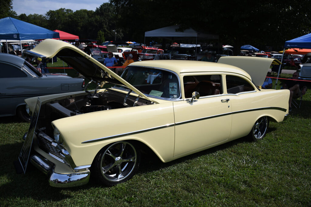 Front three-quarter view of a lowered pastel yellow 1956 Chevrolet two-door post sedan with custom billet wheels and open hood on the show field.