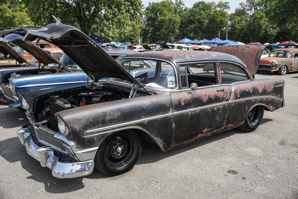 Front quarter shot of a weathered patina black 1956 Chevrolet two-door post car with black steel wheels and open hood parked at an outdoor car show.