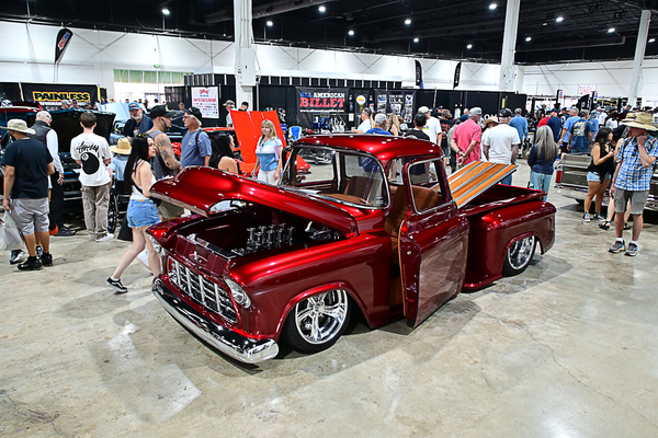 Front three-quarter view of a custom candy apple red 1955 Chevrolet pickup truck with open hood, doors, and tilt bed on display indoors at the Goodguys Pleasanton West Coast Nationals.