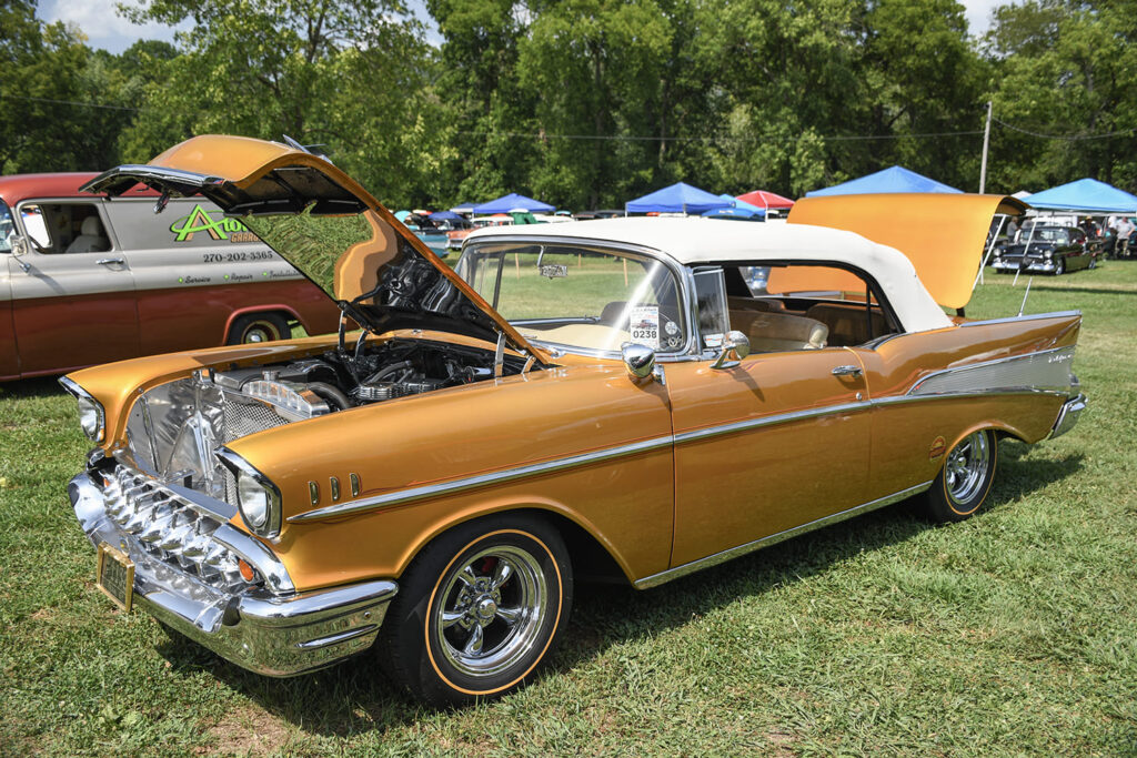 Front three-quarter view of an Inca Gold 1957 Chevrolet Bel Air convertible with a white soft top, tooth grille, and five spoke Torq-Thrust II wheels.
