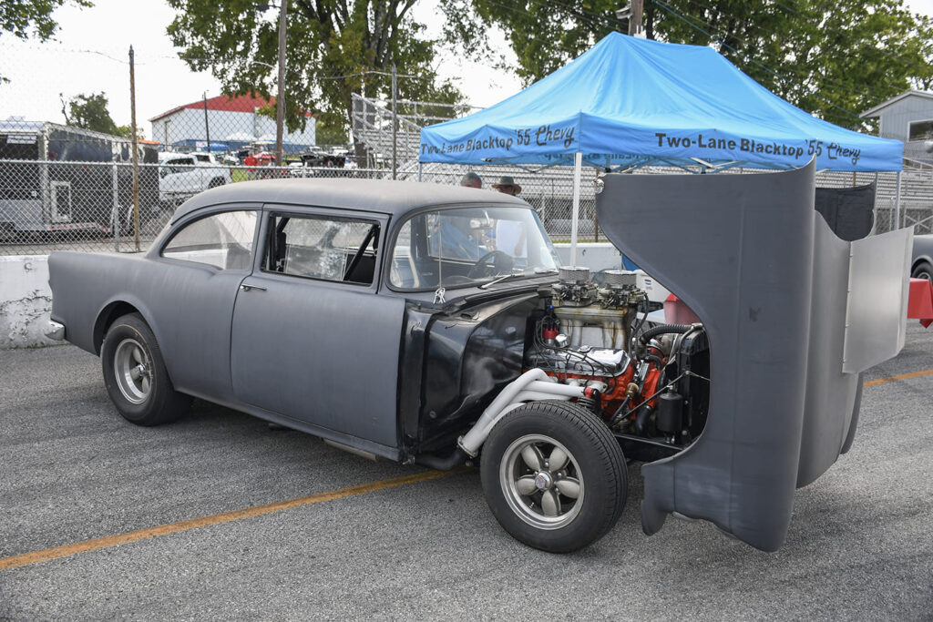 Matte gray primer 1955 Chevrolet two-door sedan gasser with a tilt front-end open, tunnel ram intake, and classic American Racing 200S wheels.
