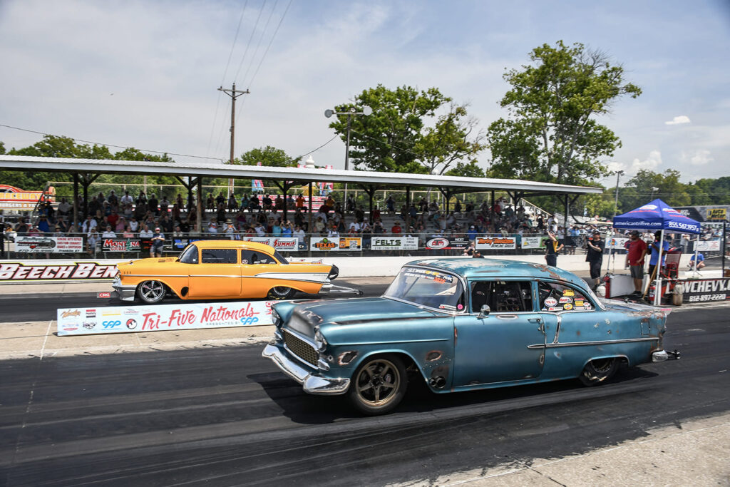 Side-by-side drag race between a weathered blue 1955 Chevy sedan and a custom yellow 1957 Chevy on the Beech Bend Raceway track.
