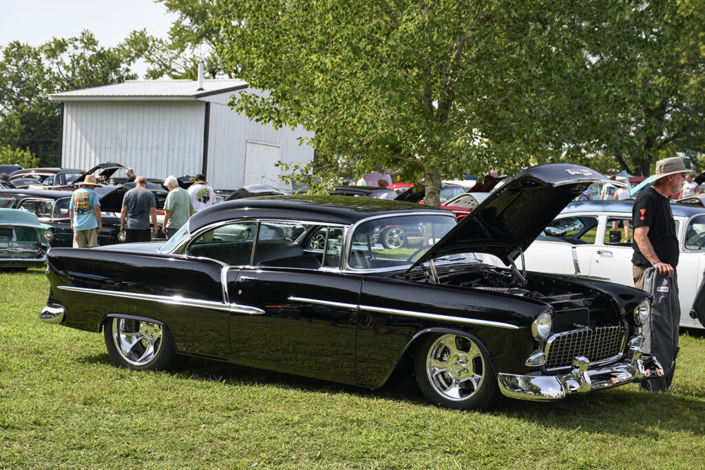 Side three-quarter profile of a deep gloss black 1955 Chevrolet Bel Air hardtop restomod with custom ET Mag-style wheels on the show grass.