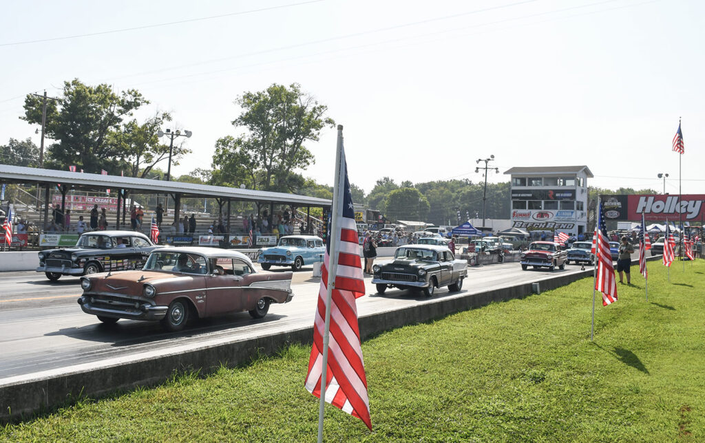 Wide-angle view of vintage 1955–1957 Chevrolets cruising down the drag strip past American flags and grandstands at the Tri-Five Nationals.