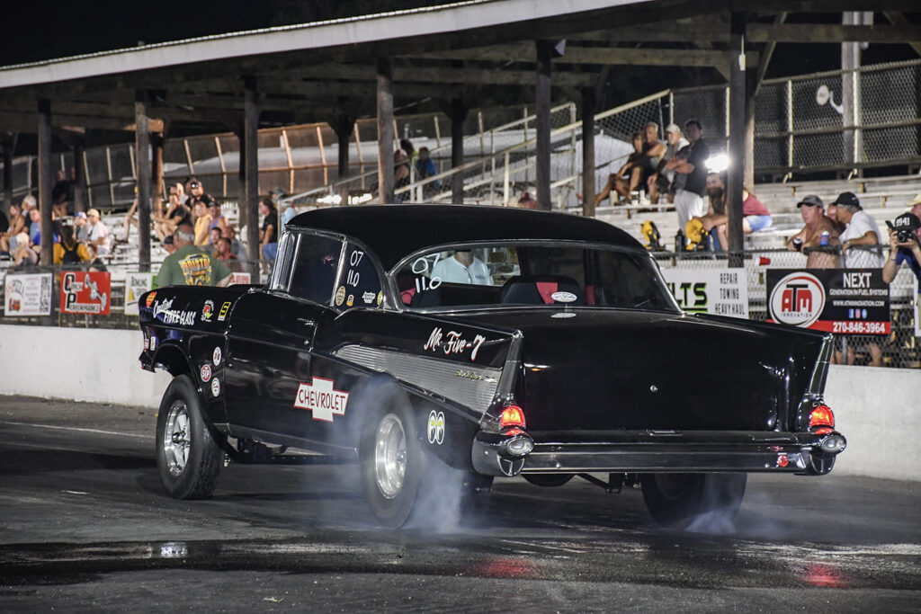 Rear three-quarter action shot of the black "Mr. Five-7" 1957 Chevrolet drag car smoking its tires under the lights at the drag strip.