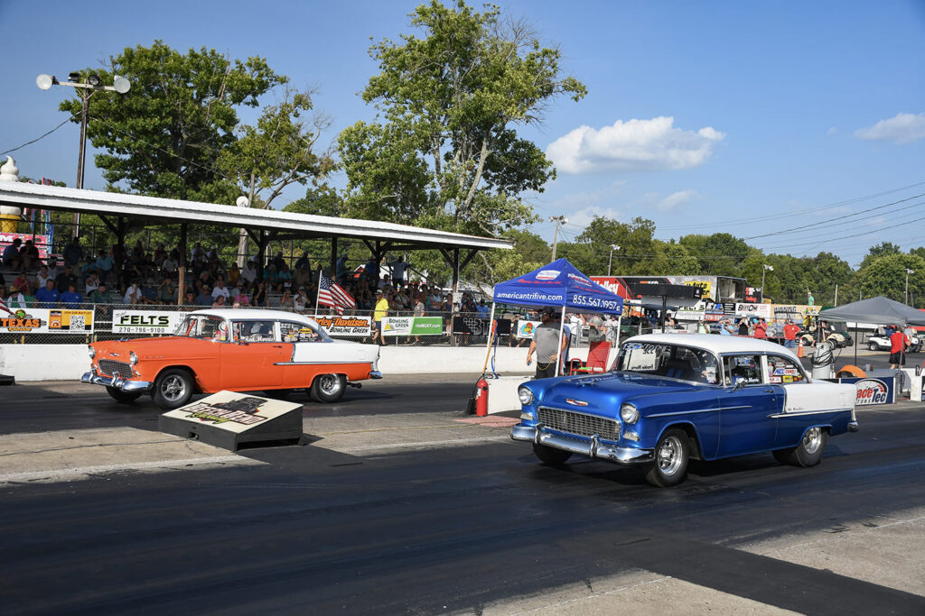 Blue 1955 Chevrolet sedan racing alongside an orange and white 1955 Chevy off the line at the Tri-Five Nationals drag strip.