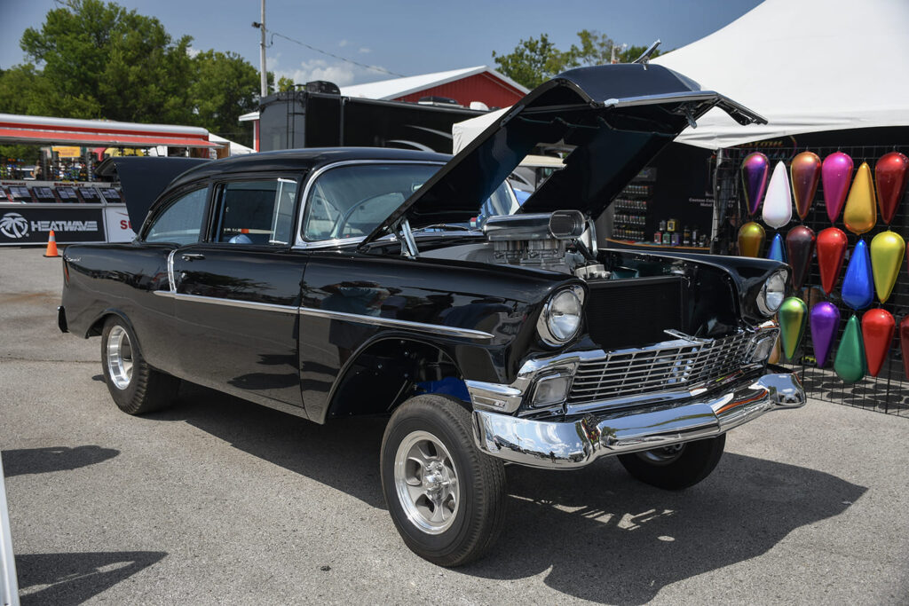Front three-quarter view of a black 1956 Chevrolet two-door post gasser with a straight front axle, raised hood, and aluminum scoop.